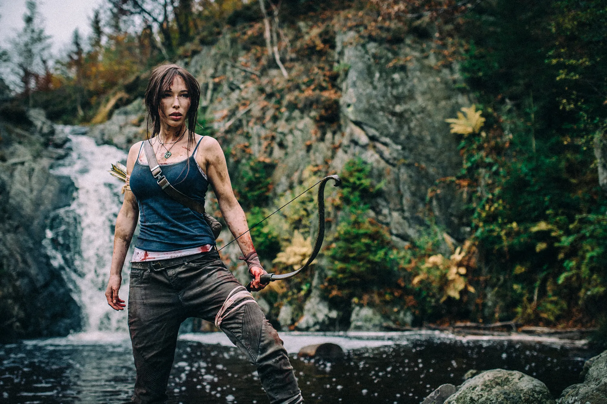 Cosplay-Porträt von Lara Croft aus Tomb Raider mit Bogen vor einer Landschaft mit Wasserfall