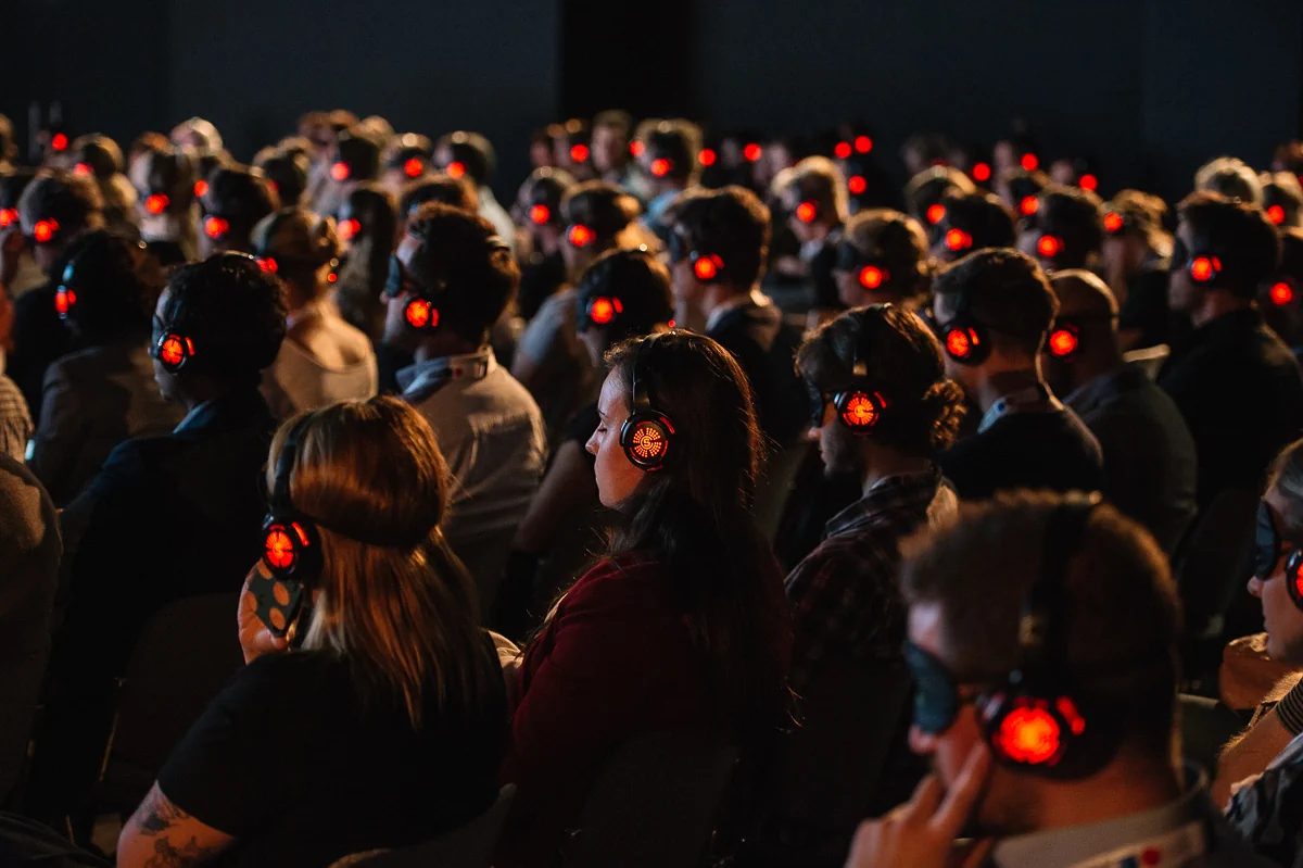 Publikum sitzt mit geschlossenen Augen und trägt rot leuchtende Kopfhörer während eines Vortrags.