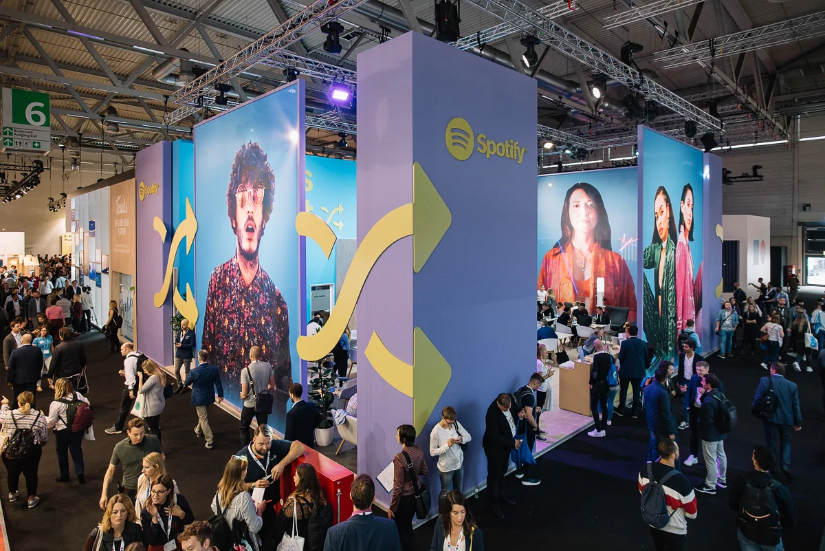 Außenansicht des Spotify-Messestands: Viele Besucher laufen durch die Gänge, im Stand selbst sind ebenfalls Personen.