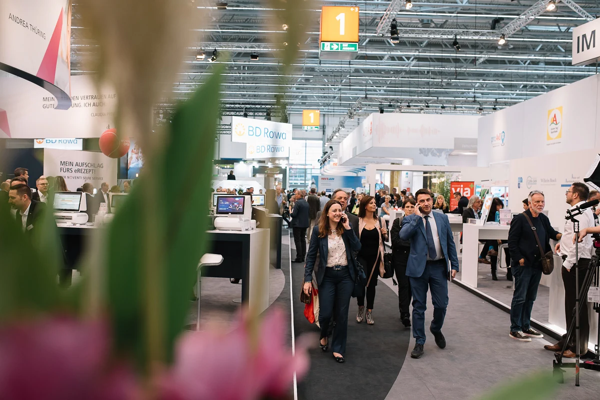 Messebesucher laufen rechts im Bild Richtung Betrachter, einige telefonieren, andere unterhalten sich im Hintergrund.