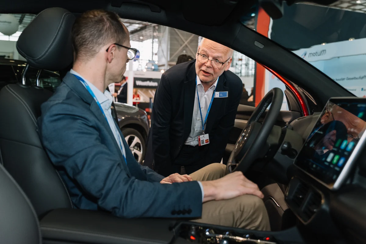 Person sitzt im Auto auf einer Messe, eine andere Person steht draußen und spricht mit ihr.